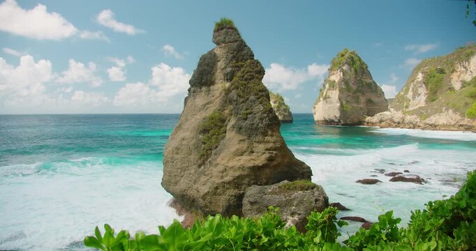 Large jagged rock pillar stands in the turquoise ocean water near a tropical coastline of Nusa Penida