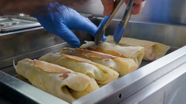 Chef using metal tongs to heat rolled wraps and burritos on a stainless steel flat top griddle in a commercial kitchen.