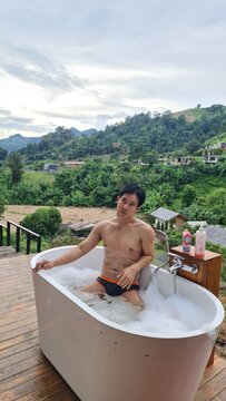Relaxed man soaking in outdoor bubble tub surrounded by tropical nature and mountains in evening time