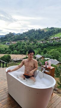 Relaxed man soaking in outdoor bubble tub surrounded by tropical nature and mountains in evening time