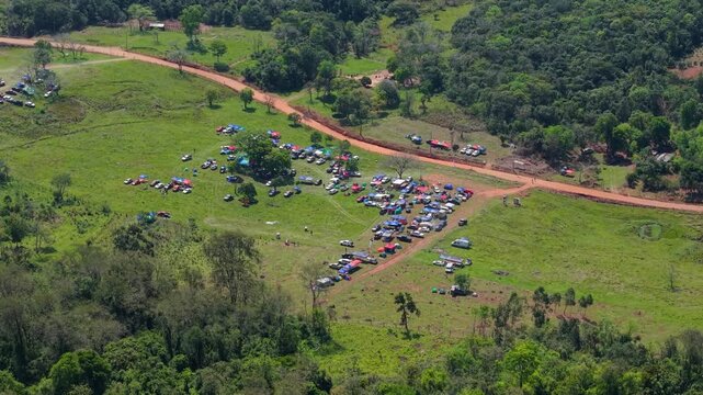 Temporary campsite with tents, parked vehicles and campers spreads across green countryside beside a rural dirt road in Nueva Alborada, Itapua, Paraguay, South America, drone establishing shot.