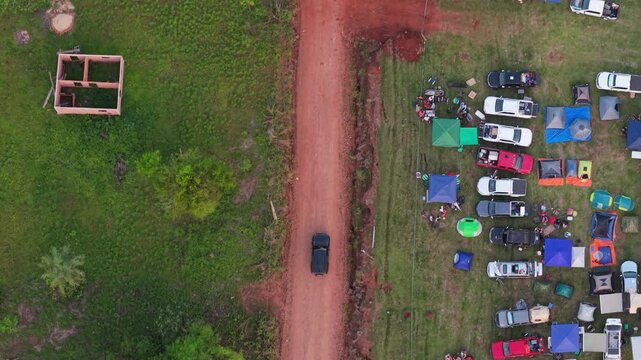 Car drives along a red dirt road beside a campsite with tents and parked vehicles in Nueva Alborada, Itapua, Paraguay, South America, top-down drone follow shot.