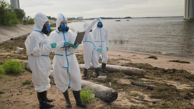 Multiethnic ecologists in hazmat suits analyzing ecological disaster data on laptop near polluted riverbank. Clip serves environmental protection campaigns and pollution research reports