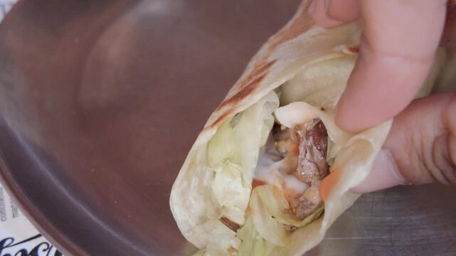 Person holding and opening fresh appetizing shawarma wrap filled with meat and vegetables.