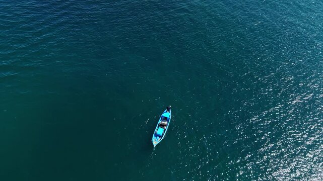 A small boat is seen floating on calm water during the day. The boat moves slowly through clear blue waves. Sunlight reflects off the surface creating a shimmering effect.