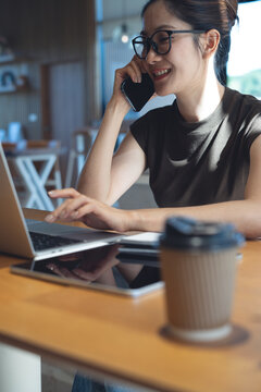 Smiling businesswoman talking on smartphone while using laptop in cafe or home office, remote work, online business, digital nomad lifestyle concept. vertical