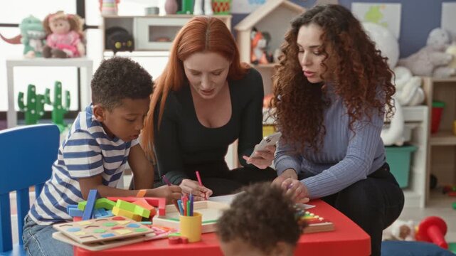 Women help boy draw with pencil and scissors at a colorful preschool play table inside a childcare building; nurturing learning focus.