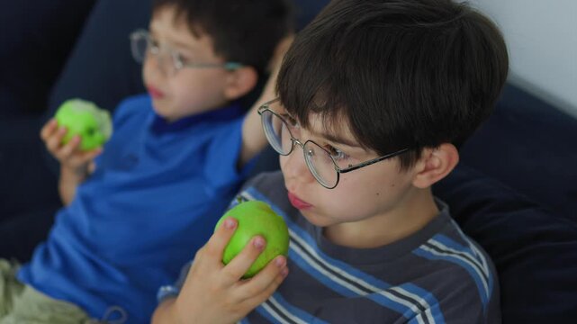 cute young boy with glasses eating a fresh green apple. Healthy childhood nutrition, daily vitamin snack, and casual indoor lifestyle portrait of a kid enjoying organic fruit.