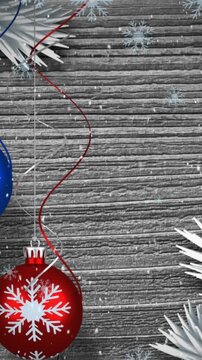 Animation of snow falling over hanging bauble decorations against wooden grey background