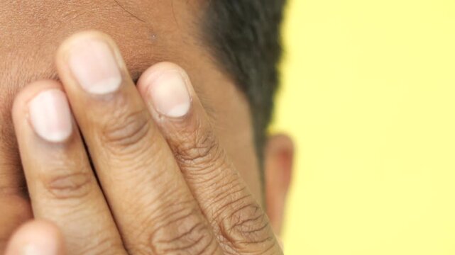 A close up view of an unrecognizable man's hands rubbing his eyes against a yellow background, tired person, eye strain, feeling sleepy