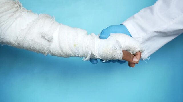 Doctor wearing blue gloves holding hand of patient with bandaged arm against blue background in studio