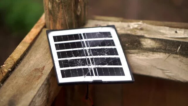 A small, cracked portable solar panel is placed on top of an outdoor wooden building frame.