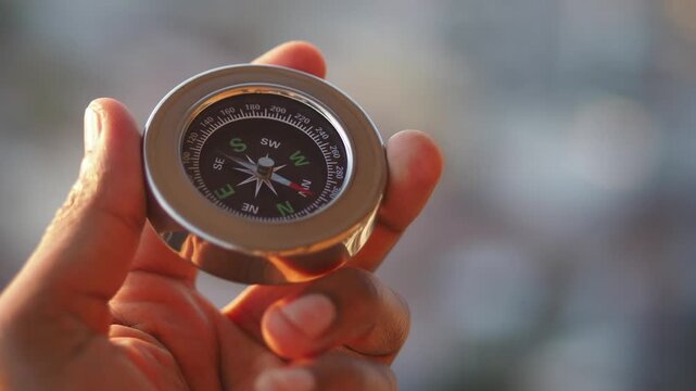 Close up of person hand holding a metal compass, needle is rotating in search of direction and finally points north with blurred background showing cityscape