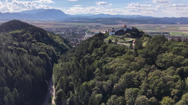 RASNOV, ROMANIA - SEPTEMBER 4, 2026: Cinematic aerial view of the medieval Rasnov Citadel seen from a distance, surrounded by lush green forests on the hills of Transylvania