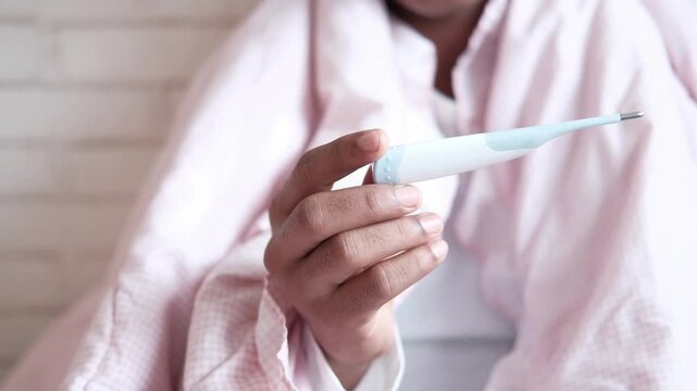 Woman holding digital thermometer under blanket while feeling sick with fever in bedroom