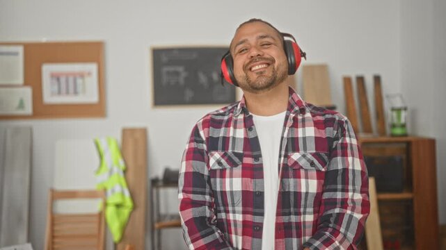 Man wearing red earmuffs smiles in a wood filled studio with planks and a high visibility vest visible; pride craft.