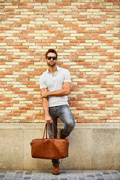 Fashion, man and portrait for travel by brick wall with duffle bag for luggage, space and outdoor. Male tourist, building and trendy style in city with smart casual outfit, urban trip and confidence