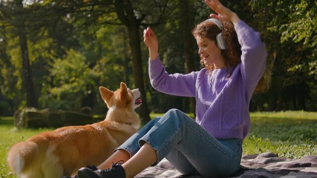 Laughing young woman in headphones playing train welsh corgi dog in city park outdoors happy female girl handler animal trainer exercising puppy little doggie waiting command feeding treats in nature