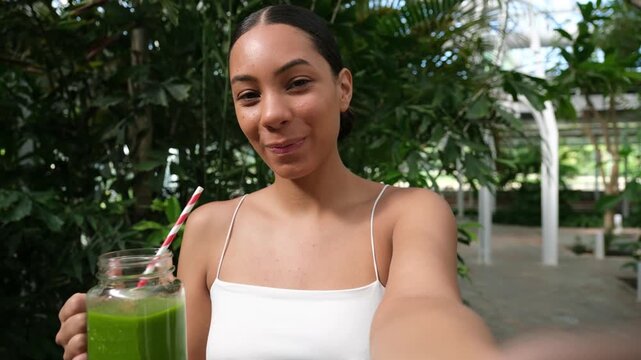 Happy woman filming herself on camera enjoying a healthy green juice outdoors