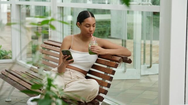 Smiling young woman drinking a healthy green smoothie while using her phone on a bench
