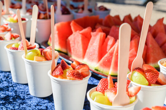 Individual cups of fresh fruit salad with wooden spoons arranged on a table during an outdoor picnic. The healthy fruit portions are prepared for casual social dining in a relaxed outdoor setting