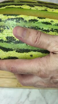 Male hands holding a large knife and cutting a fresh, ripe melon in half lengthwise on a wooden cutting board in the kitchen.