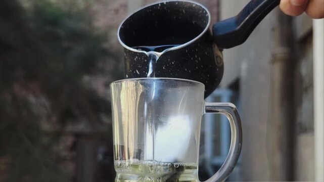 Close-up of hot water being poured from a small black pot into a clear glass mug containing a green tea bag, preparing a hot herbal beverage outdoors in warm natural light.