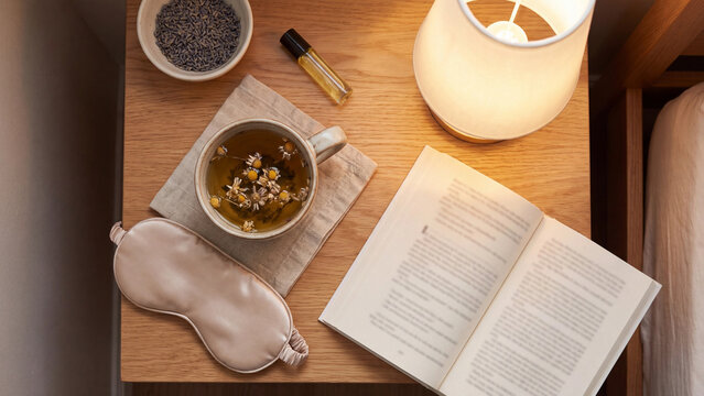 Bedside table illuminated by a warm lamp with an open book, herbal tea, sleep mask, and lavender.