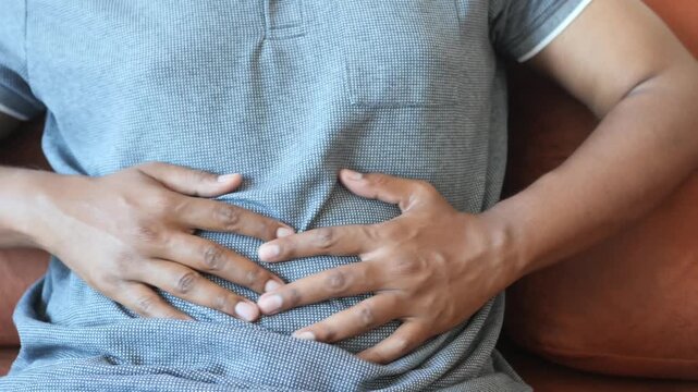 A man wearing a grey polo shirt is seen from the chest up, rubbing his stomach with both hands, indicating stomach pain or indigestion as he sits on a brown couch