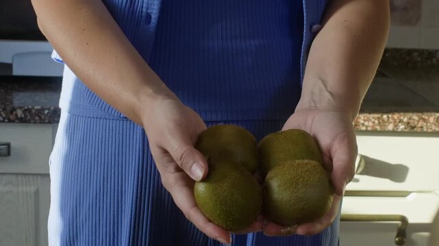 Woman in blue dress displays fresh kiwi fruits in cupped hands indoors, static camera captures natural product handling, warm kitchen setting, raw food concept.