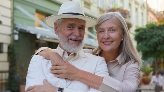 Happy Caucasian elderly couple husband and wife hugging looking at camera on city street loving senior family people enjoying romantic retirement lifestyle travel vacations happiness marriage outdoors
