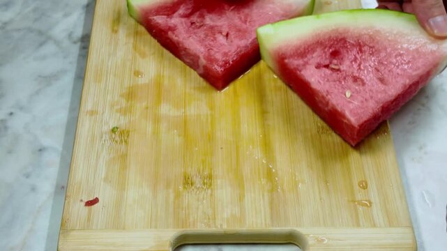 Person's hands using a kitchen knife to cut the green rind off fresh, juicy red watermelon slices on a wooden cutting board