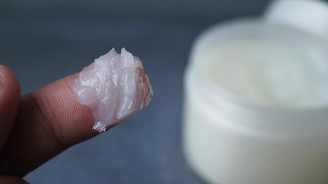 Close up of petroleum jelly on a fingertip near a white jar, skincare and beauty product