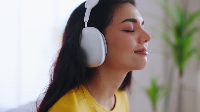 Asian woman meditates and practices Zen meditation at home, focusing on breathing exercises and seeking spiritual peace in her living room. She sits on the sofa, listening podcast to headphones