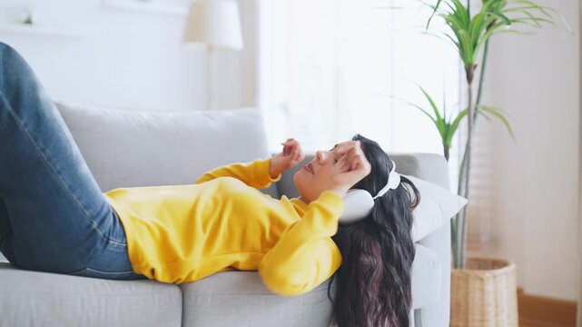 Asian woman is listening to music or a podcast through headphones to relax in the living room. Young lady is enjoying lounging on the sofa at home. Meditation for mental health and wellbeing