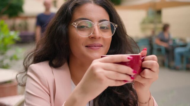 Happy young Indian woman holding warm cup of coffee sitting outside city cafe smiling relaxed Arabian ethnic female girl lady close up portrait client customer enjoying drink tea shop town restaurant