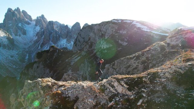 Static drone view captures male hiker crossing rocky alpine terrain near Tre Cime di Lavaredo, with rugged peaks, snow patches and bright evening backlight across Dolomites, Italy