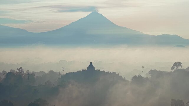 Aerial sunrise view of Borobudur temple silhouette with Merapi volcano in background during morning mist.