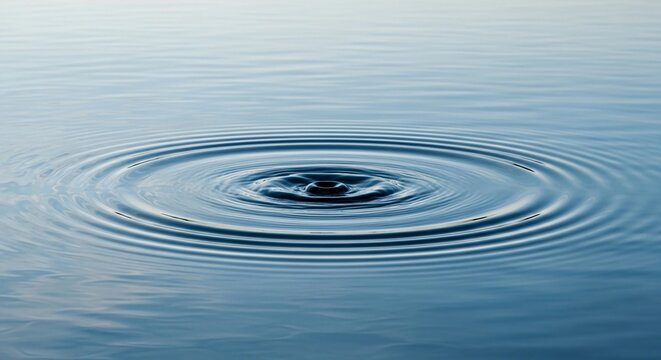 A single water droplet creates expanding ripples across a calm, blue liquid surface