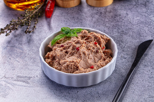 Liver pate in white ceramic bowl with basil and pink peppercorns on gray stone surface