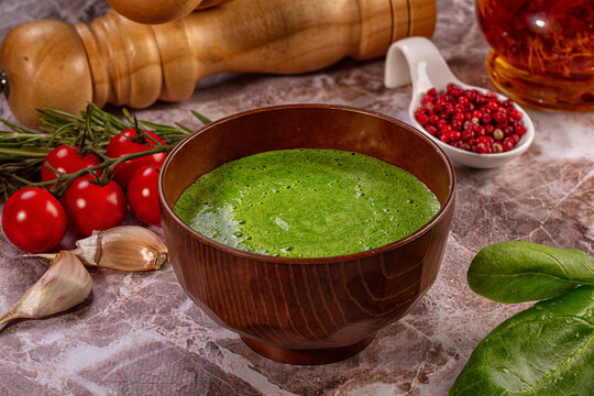 Green spinach cream soup in wooden bowl with cherry tomatoes, garlic, and peppercorns on marble surface