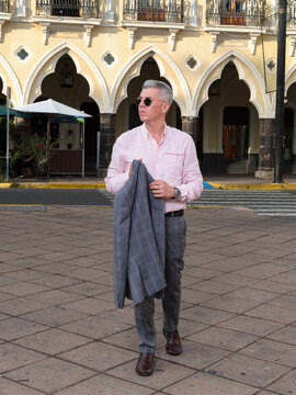 Stylish Mature Man in Pink Shirt and Sunglasses Holding a Gray Jacket in Historic Plaza q