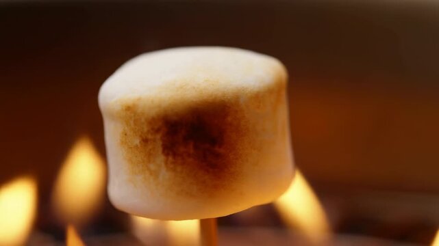 Marshmallow on wooden skewer slowly toasts over flickering flames as its white surface turns golden brown and develops a dark roasted patch in warm close-up view