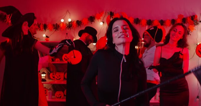 Young happy woman in black devil costume dancing with friends at Halloween party holding scythe in hands. People group having fun celebrating holiday at home. Celebration concept. 4k video.