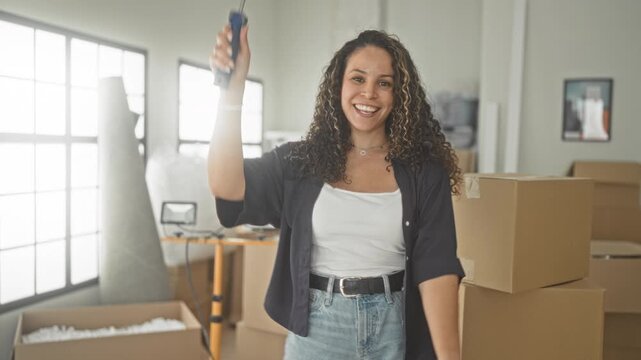 Woman holding a paint roller and raising her arm among packed moving boxes in a bright studio; joyful fresh start.