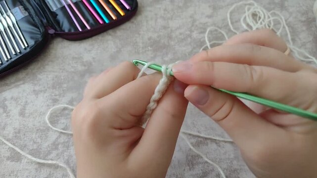 Hands crocheting a second row of single stitches into a white yarn chain using a green aluminum hook. Horizontal stock footage for craft tutorials, DIY knitting, and creative hobby vlogs.