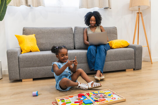 African american mother using a laptop while her young daughter plays with learning toys on the living room floor at home