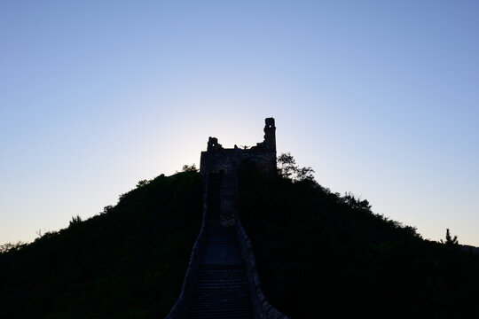 Dark silhouette of a broken ancient watchtower on the Jinshanling Great Wall atop a green hill, set against a soft pale twilight sky before sunrise.
