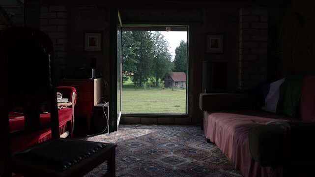 Dim rustic room with red upholstered furniture and patterned carpet framing a bright view through an open doorway toward grass, trees, and a small wooden outbuilding