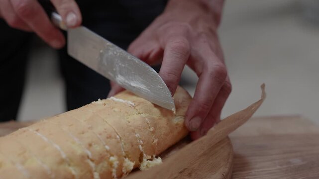 Close-up of hands cutting a homemade sponge roll cake into even slices on parchment paper over a wooden serving board, with cream filling visible between layers